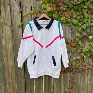 Puede incluir: Chaqueta blanca vintage con cuello azul marino y detalles en verde azulado y rosa intenso. La chaqueta tiene cierre de botones en la parte delantera y dos bolsillos azul marino. Cuelga de una percha de madera contra una valla de madera.