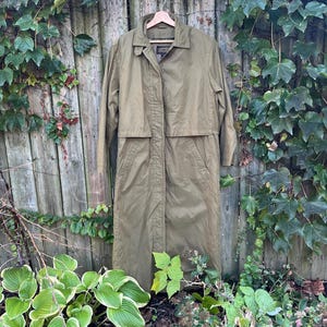 May include: A long, olive green trench coat hanging on a wooden fence. The coat has a collared neckline, a button-down front, and a mid-thigh length. The coat is made of a lightweight material, perfect for spring or fall weather.