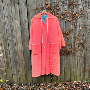 May include: Coral quilted robe with white trim and a turquoise lining, hanging on a wooden hanger. The robe has a diamond pattern, pockets, and white rope ties. The robe is displayed against a weathered wooden fence.