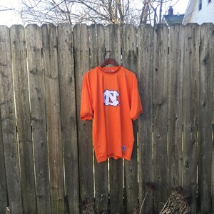 K&ouml;nnte beinhalten: Orangefarbenes T-Shirt mit einem schwarzen und wei&szlig;en "N"-Logo auf der Vorderseite. Das Shirt hat einen schwarzen Saum an Hals und &Auml;rmeln.