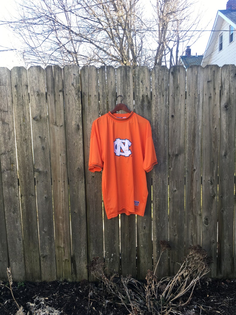 K&ouml;nnte beinhalten: Orangefarbenes T-Shirt mit einem schwarzen und wei&szlig;en "N"-Logo auf der Vorderseite. Das Shirt hat einen schwarzen Saum an Hals und &Auml;rmeln.