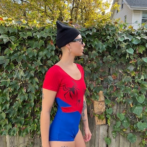 Pu&ograve; includere: Un costume da bagno intero rosso e blu con una grafica di un ragno nero. Il costume da bagno ha maniche corte e una scollatura rotonda. La modella indossa un berretto nero e occhiali da sole.
