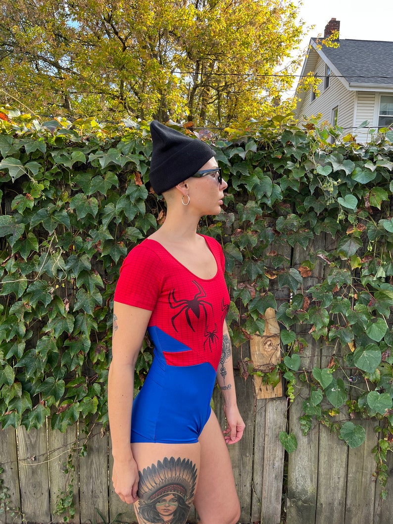 Pu&ograve; includere: Un costume da bagno intero rosso e blu con una grafica di un ragno nero. Il costume da bagno ha maniche corte e una scollatura rotonda. La modella indossa un berretto nero e occhiali da sole.