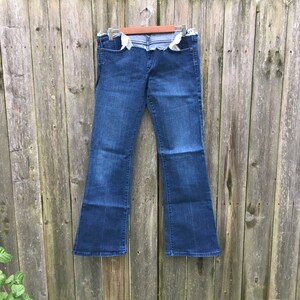 May include: Dark wash denim jeans with a flared leg, hanging on a wooden fence. The jeans have a distressed, unfinished waistline and pockets. The jeans are a classic style, suitable for casual wear.