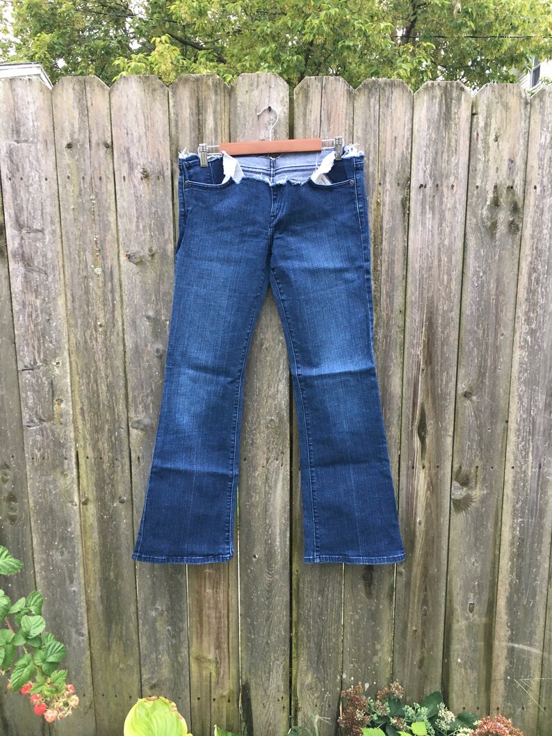 May include: Dark wash denim jeans with a flared leg, hanging on a wooden fence. The jeans have a distressed, unfinished waistline and pockets. The jeans are a classic style, suitable for casual wear.