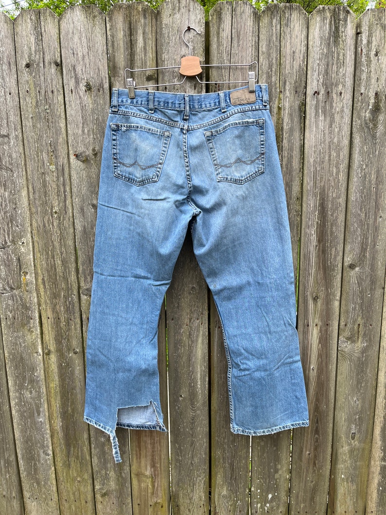 May include: A pair of light blue denim jeans with a distressed hem. The jeans have a straight leg and are hanging on a wooden hanger.