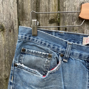 May include: A pair of blue denim jeans with a red and white plaid lining peeking out from a ripped pocket.