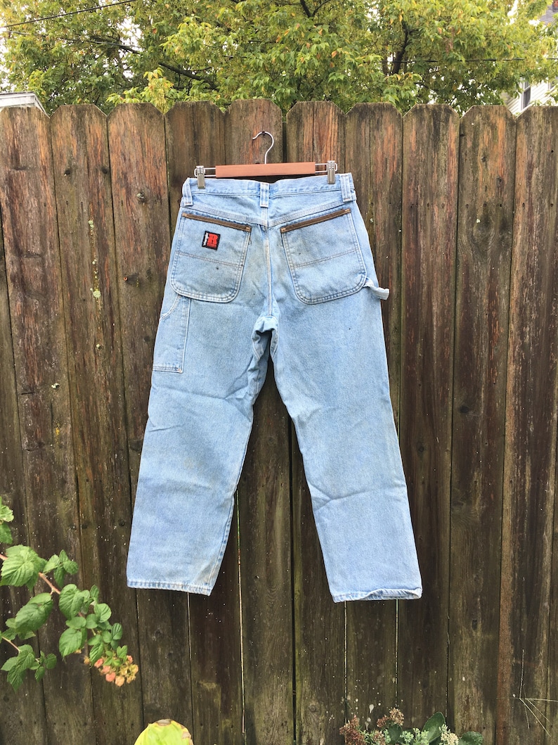 May include: Light blue denim carpenter jeans with a brown leather patch on the back pocket. The patch has a red and white logo.