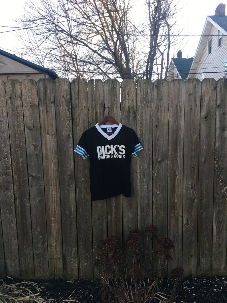 May include: Black athletic t-shirt with white collar and sleeve stripes. The shirt features the text "DICK'S SPORTING GOODS" in white block letters. The shirt is hanging on a black hanger against a wooden fence.