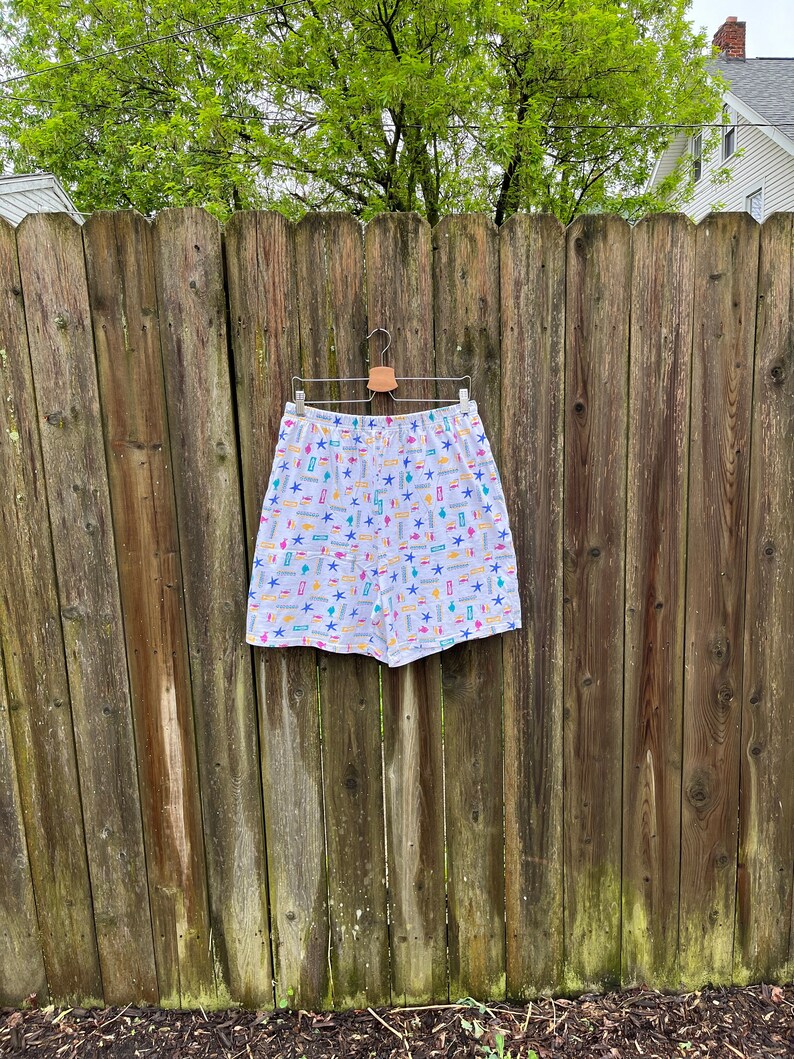 May include: White shorts with a colourful pattern of stars, geometric shapes, and lines. The shorts are hanging on a wooden hanger against a wooden fence.