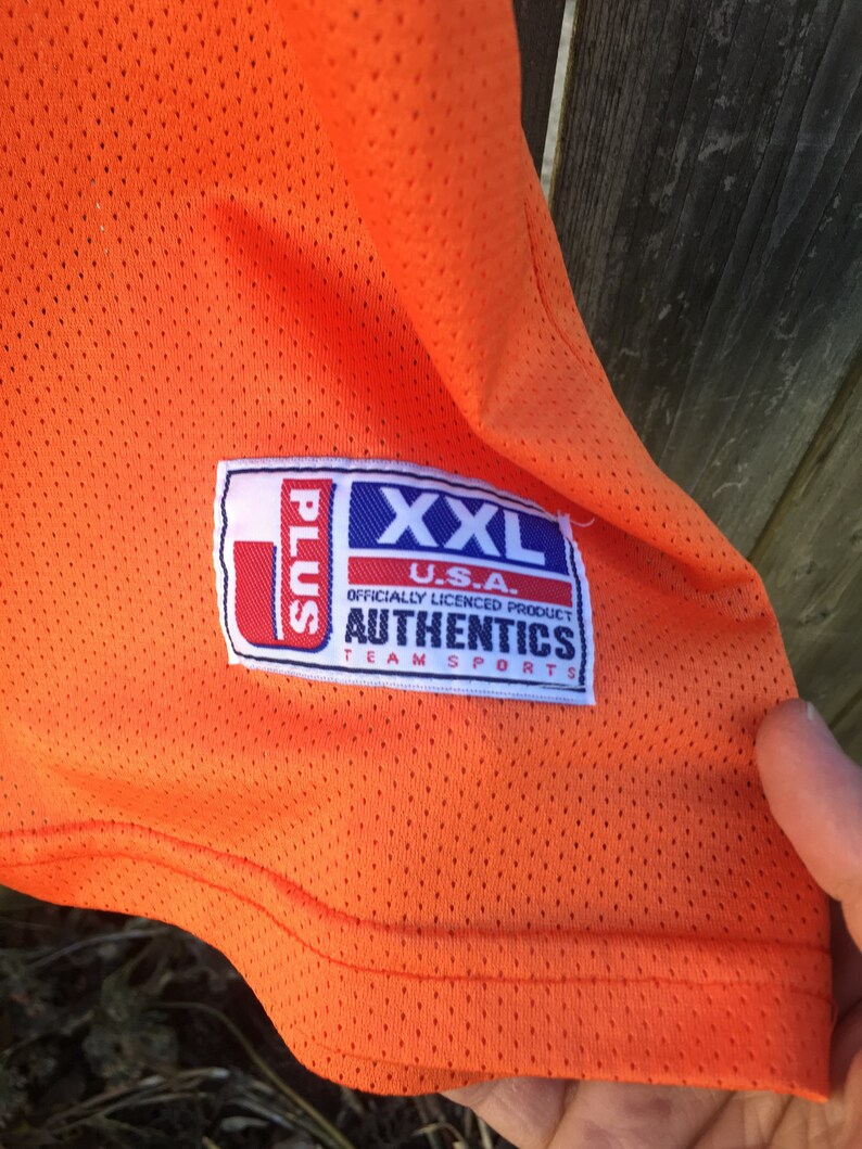 K&ouml;nnte beinhalten: Orangefarbenes Mesh-Trikot mit einem wei&szlig;en und blauen Etikett mit der Aufschrift "U.S.A. XXL Plus Offiziell lizenziertes Produkt Authentics Team Sports".