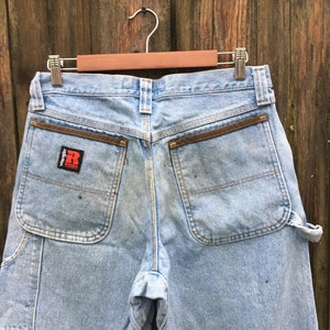 May include: A pair of light blue denim carpenter jeans with brown leather accents on the back pockets. The jeans have a Wrangler logo patch on the back right pocket.