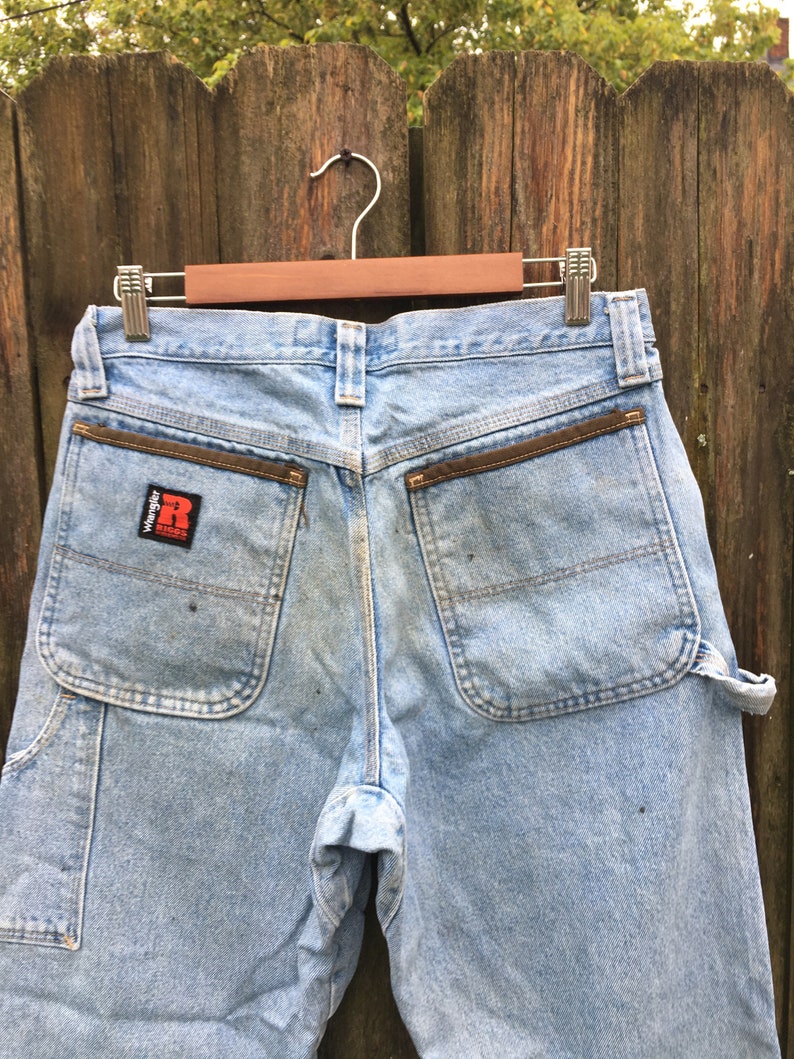 May include: A pair of light blue denim carpenter jeans with brown leather accents on the back pockets. The jeans have a Wrangler logo patch on the back right pocket.