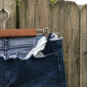 May include: Close-up of denim shorts with a distressed, frayed hem. The shorts are a medium wash blue with a lighter blue panel at the waist. The pocket lining is white. The shorts are hanging on a wooden hanger against a wooden fence.
