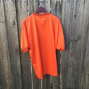 K&ouml;nnte beinhalten: Orangefarbenes T-Shirt mit kurzen &Auml;rmeln und schwarzem Besatz an Kragen und &Auml;rmeln. Das Shirt h&auml;ngt an einem Holzb&uuml;gel vor einem Holzzaun.