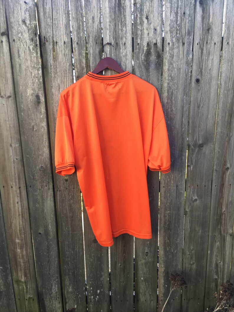 K&ouml;nnte beinhalten: Orangefarbenes T-Shirt mit kurzen &Auml;rmeln und schwarzem Besatz an Kragen und &Auml;rmeln. Das Shirt h&auml;ngt an einem Holzb&uuml;gel vor einem Holzzaun.