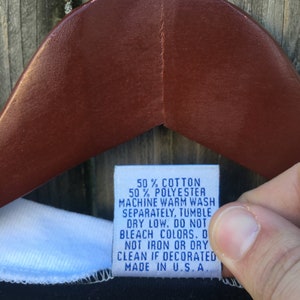 May include: A close-up of a clothing tag with the text: 50% COTTON, 50% POLYESTER, MACHINE WARM WASH, SEPARATELY, TUMBLE DRY LOW, DO NOT BLEACH COLORS, DO NOT IRON OR DRY CLEAN IF DECORATED, MADE IN U.S.A. The tag is held by a person.