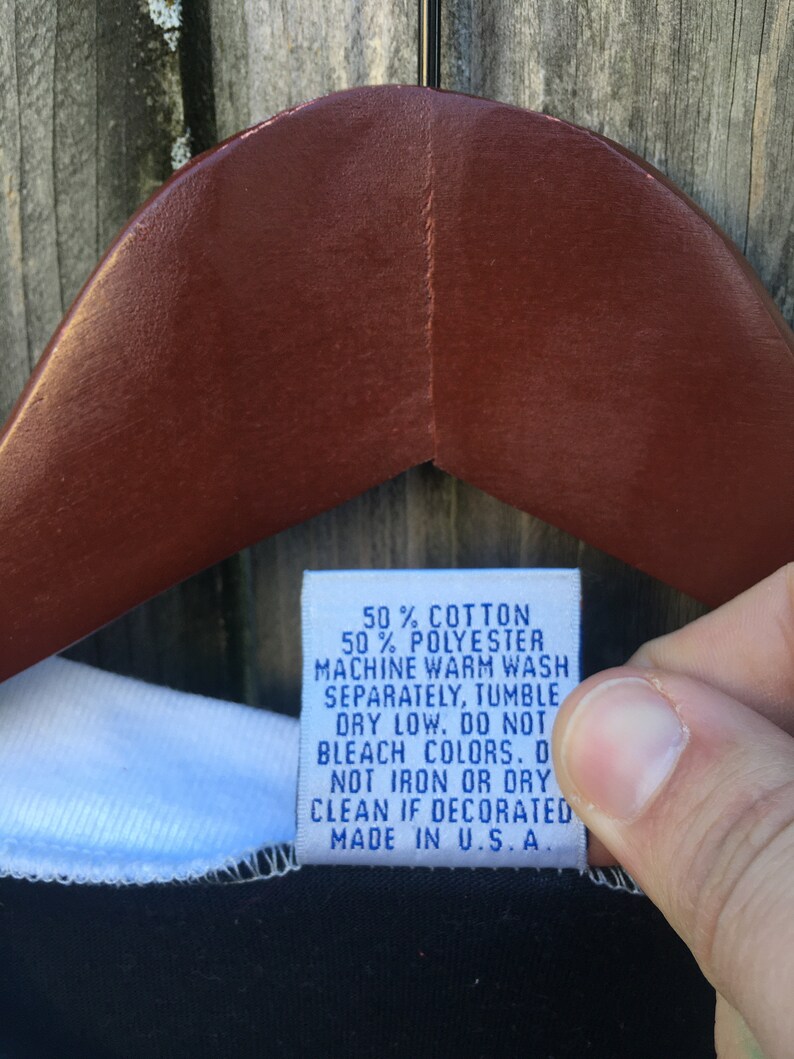 May include: A close-up of a clothing tag with the text: 50% COTTON, 50% POLYESTER, MACHINE WARM WASH, SEPARATELY, TUMBLE DRY LOW, DO NOT BLEACH COLORS, DO NOT IRON OR DRY CLEAN IF DECORATED, MADE IN U.S.A. The tag is held by a person.