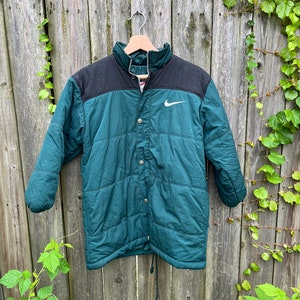 May include: A green and black puffer jacket with a white Nike swoosh logo on the chest. The jacket has a snap button closure and a drawstring at the bottom.