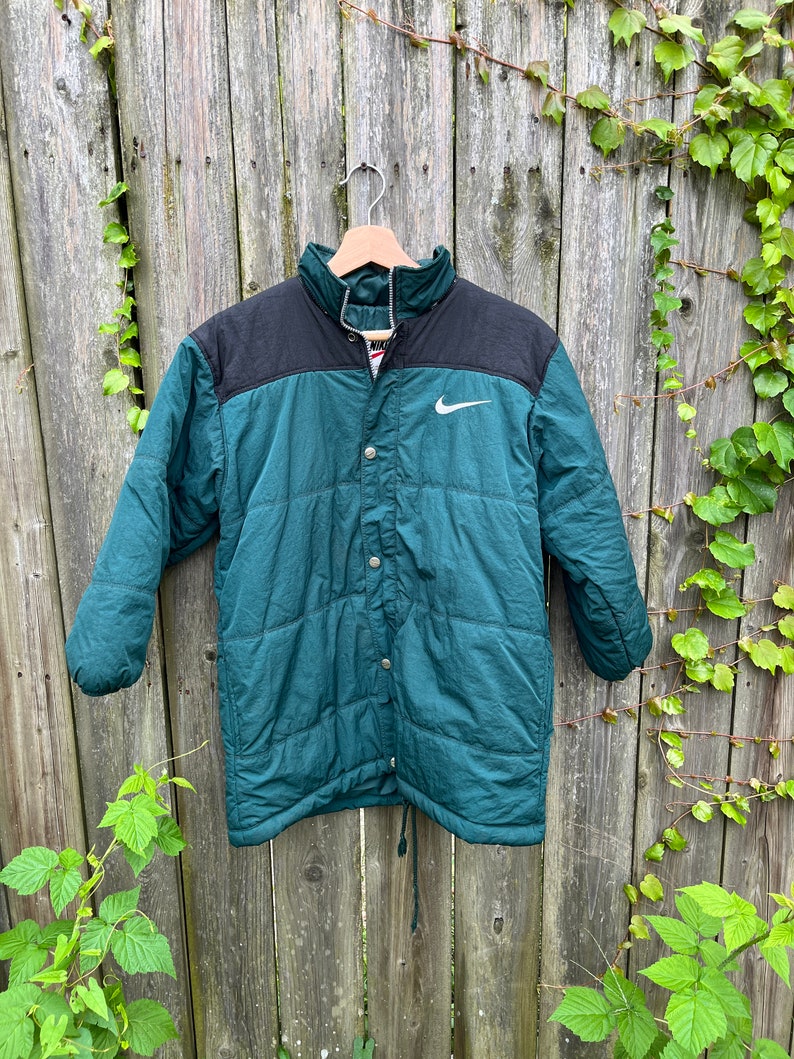 May include: A green and black puffer jacket with a white Nike swoosh logo on the chest. The jacket has a snap button closure and a drawstring at the bottom.