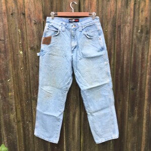 May include: Light blue denim carpenter jeans with a brown leather patch on the right pocket. The jeans are hanging on a wooden hanger against a wooden fence.