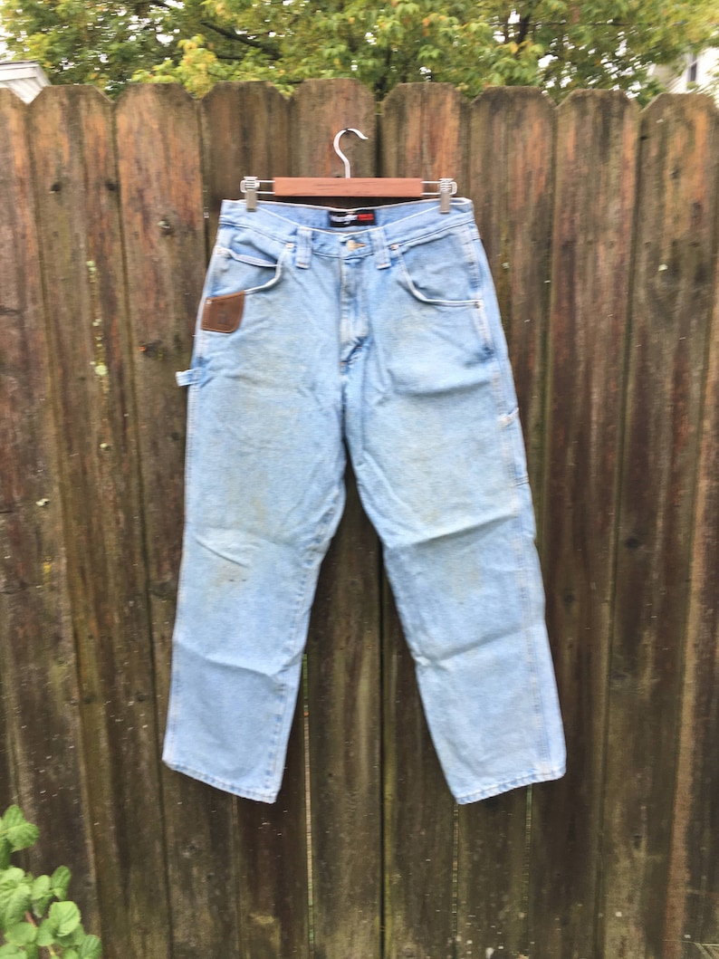 May include: Light blue denim carpenter jeans with a brown leather patch on the right pocket. The jeans are hanging on a wooden hanger against a wooden fence.