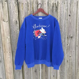 May include: A royal blue sweatshirt with a white angel graphic and the word "Believe" in white cursive. The angel is wearing a red dress and white wings.