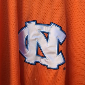 K&ouml;nnte beinhalten: Orangefarbenes Trikot mit einem wei&szlig;en und blauen "NC"-Logo.