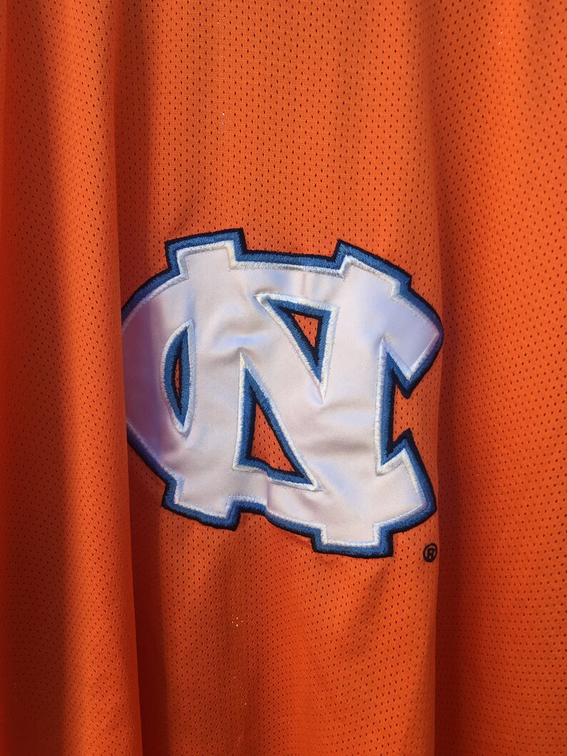 K&ouml;nnte beinhalten: Orangefarbenes Trikot mit einem wei&szlig;en und blauen "NC"-Logo.