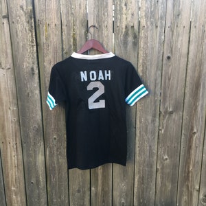 May include: Black jersey-style t-shirt with white and teal stripes on the sleeves. The name "NOAH" and the number "2" are printed on the back in silver. The shirt is hanging on a wooden hanger against a weathered wooden fence.