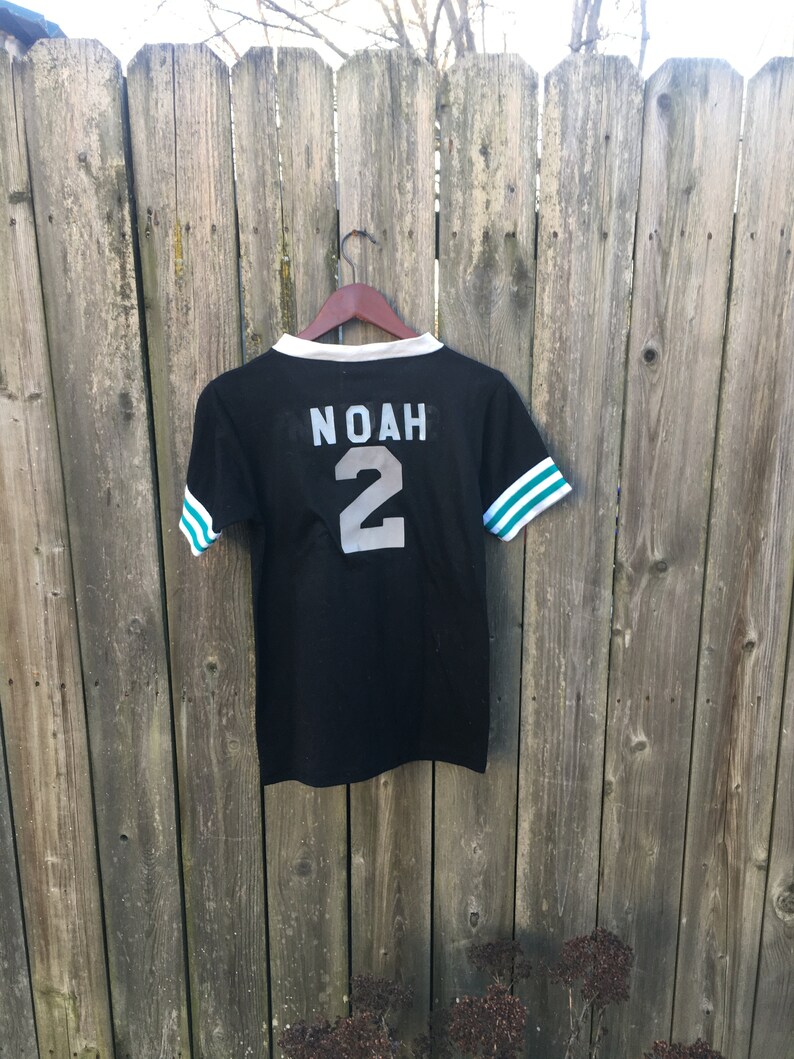 May include: Black jersey-style t-shirt with white and teal stripes on the sleeves. The name "NOAH" and the number "2" are printed on the back in silver. The shirt is hanging on a wooden hanger against a weathered wooden fence.