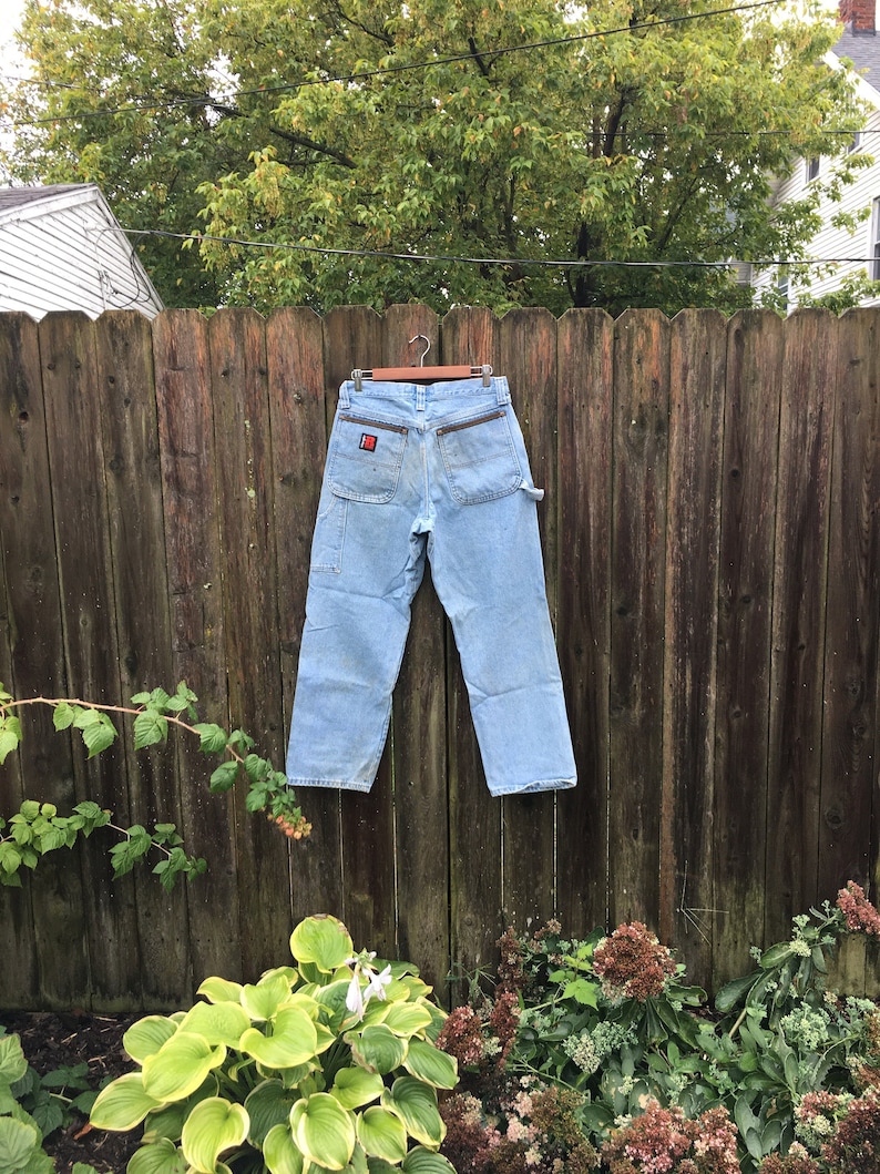 May include: A pair of light blue carpenter jeans hanging on a wooden fence. The jeans have a red patch on the back pocket with the text "Dickies" on it.