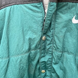 May include: A green and black nylon jacket with a zipper closure and a white Nike logo on the right side. The jacket has a buttoned flap over the zipper.