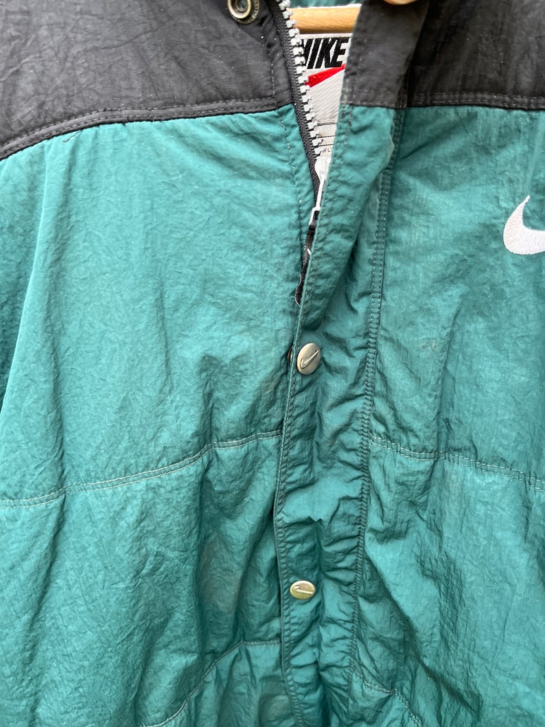 May include: A green and black nylon jacket with a zipper closure and a white Nike logo on the right side. The jacket has a buttoned flap over the zipper.