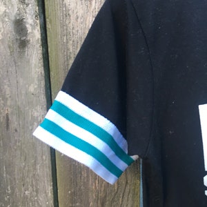 May include: Black short-sleeved shirt with white and teal striped sleeve cuffs. The shirt's sleeve is visible against a weathered wooden background. The cuffs have three teal stripes between two white stripes.