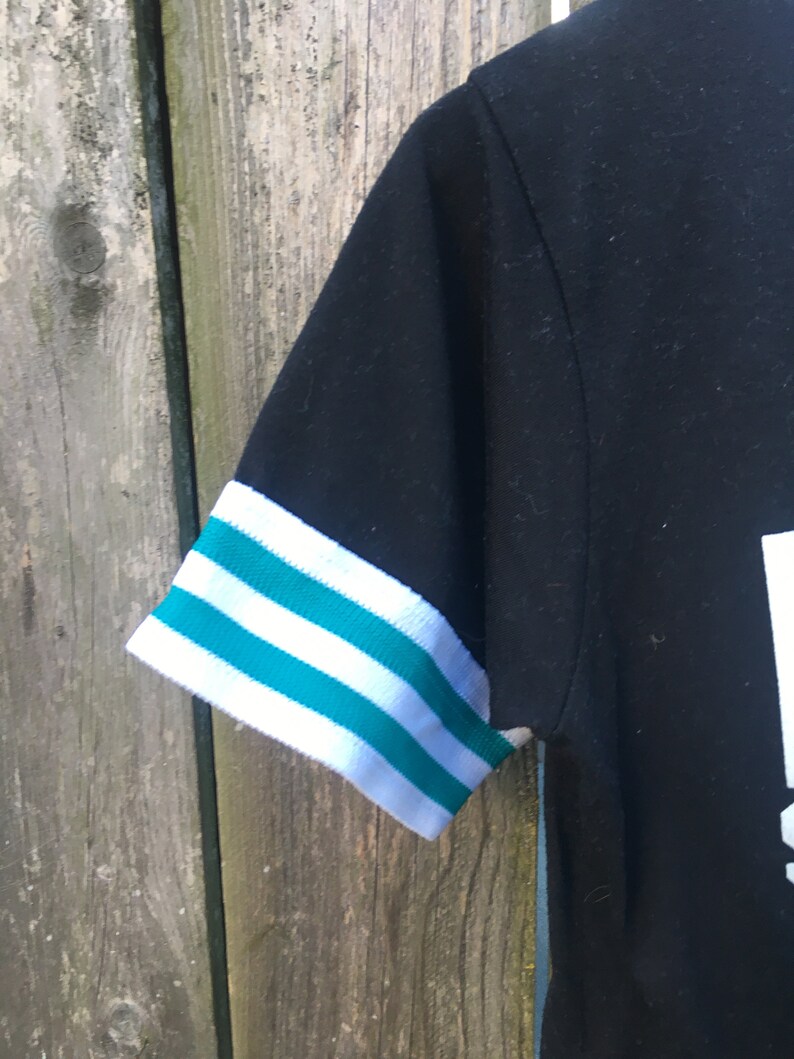 May include: Black short-sleeved shirt with white and teal striped sleeve cuffs. The shirt's sleeve is visible against a weathered wooden background. The cuffs have three teal stripes between two white stripes.