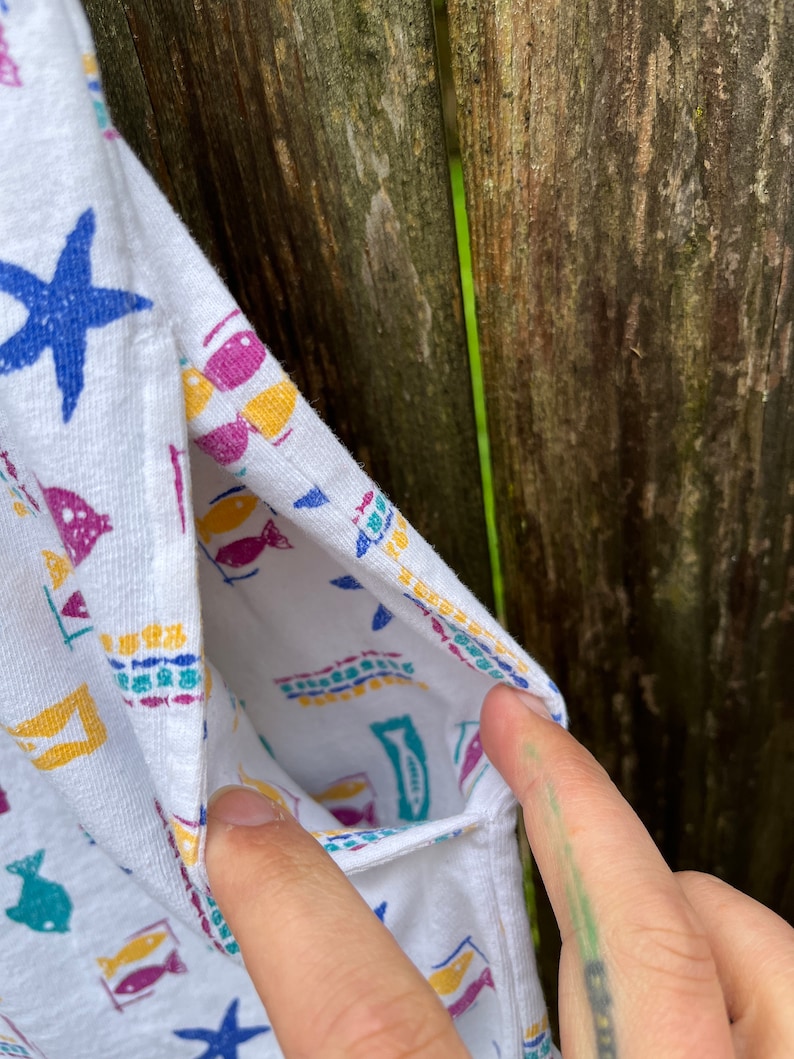 May include: White sleeveless shirt with a colourful fish and starfish pattern. The shirt has a pocket on the left side.