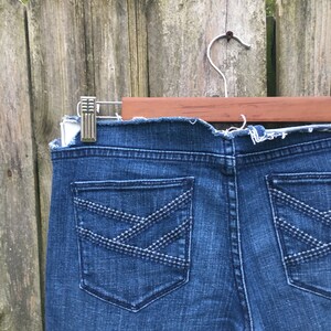 May include: Close-up of the back of blue denim jeans. The jeans have two back pockets with decorative stitching in a crisscross pattern. The jeans are hanging on a wooden hanger.