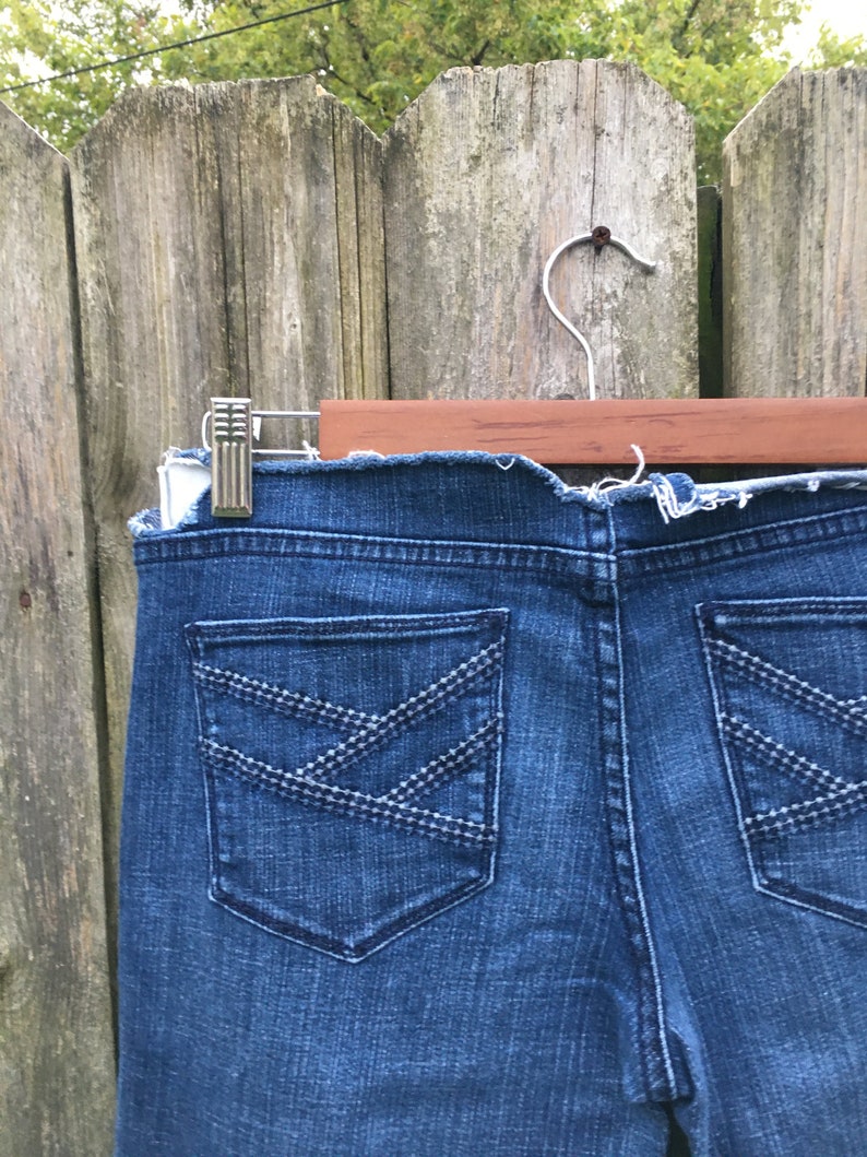 May include: Close-up of the back of blue denim jeans. The jeans have two back pockets with decorative stitching in a crisscross pattern. The jeans are hanging on a wooden hanger.