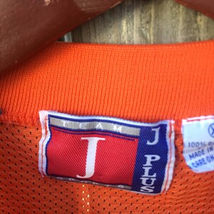 K&ouml;nnte beinhalten: Orangefarbenes Mesh-Trikot mit einem roten und blauen "J Plus Team"-Etikett. Das Etikett hat ein gro&szlig;es wei&szlig;es "J" in der Mitte.