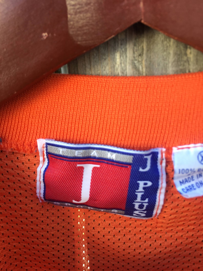 K&ouml;nnte beinhalten: Orangefarbenes Mesh-Trikot mit einem roten und blauen "J Plus Team"-Etikett. Das Etikett hat ein gro&szlig;es wei&szlig;es "J" in der Mitte.