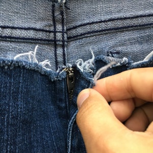 May include: Close-up of the zipper and fly of a pair of blue denim jeans. The jeans have a distressed, frayed edge along the top. A hand is reaching to zip up the jeans.