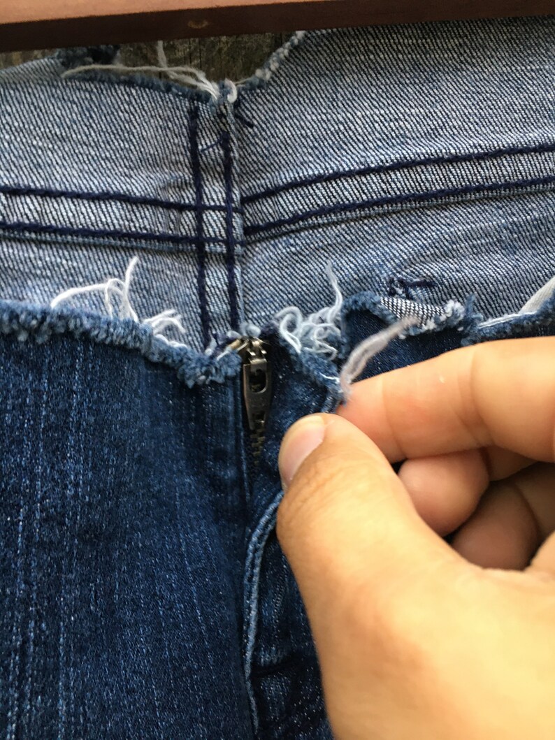 May include: Close-up of the zipper and fly of a pair of blue denim jeans. The jeans have a distressed, frayed edge along the top. A hand is reaching to zip up the jeans.