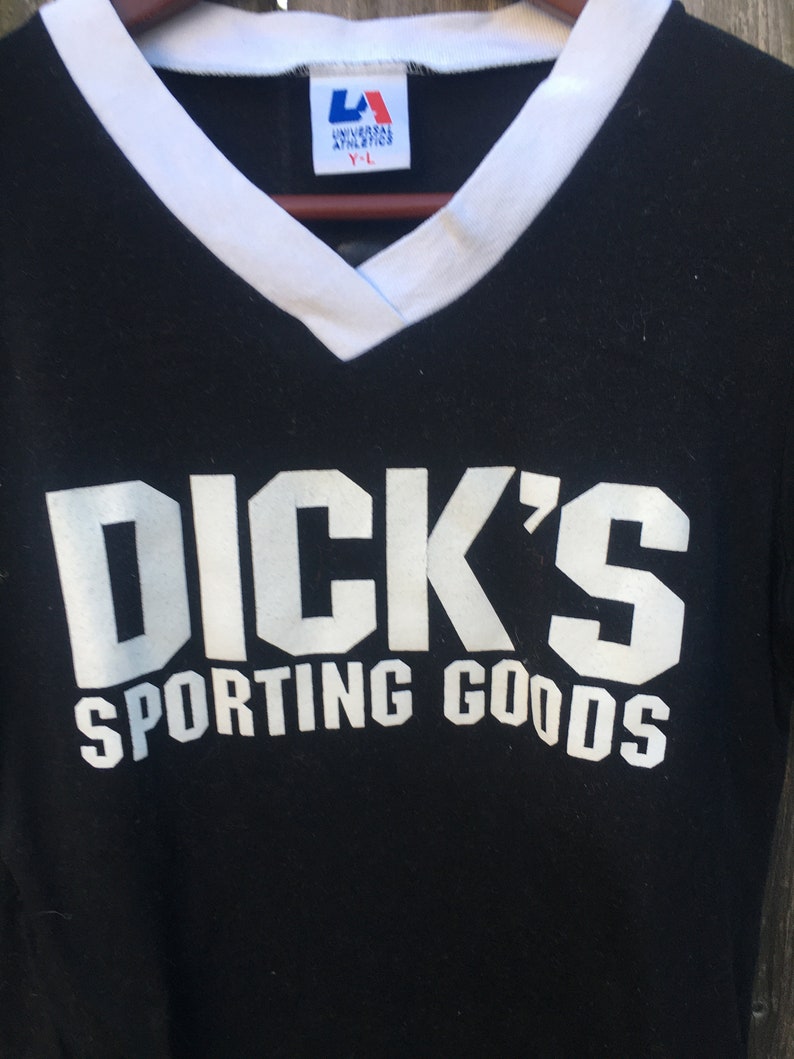 May include: Black v-neck t-shirt with white trim and the text "DICK'S SPORTING GOODS" in white block letters. The shirt has a tag that reads "UNIVERSAL ATHLETICS Y-L".