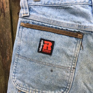 May include: Light blue denim carpenter jeans with a brown pocket flap and a red and white Wrangler Riggs Workwear patch.