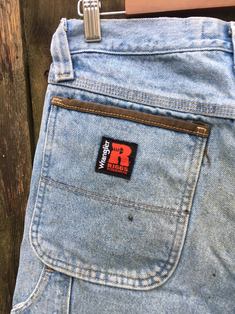May include: Light blue denim carpenter jeans with a brown pocket flap and a red and white Wrangler Riggs Workwear patch.