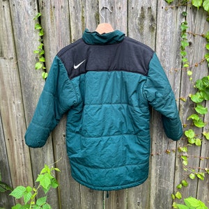 May include: A dark green and black puffer jacket with a white Nike swoosh logo on the upper left chest. The jacket has a zipper closure and a drawstring at the bottom.