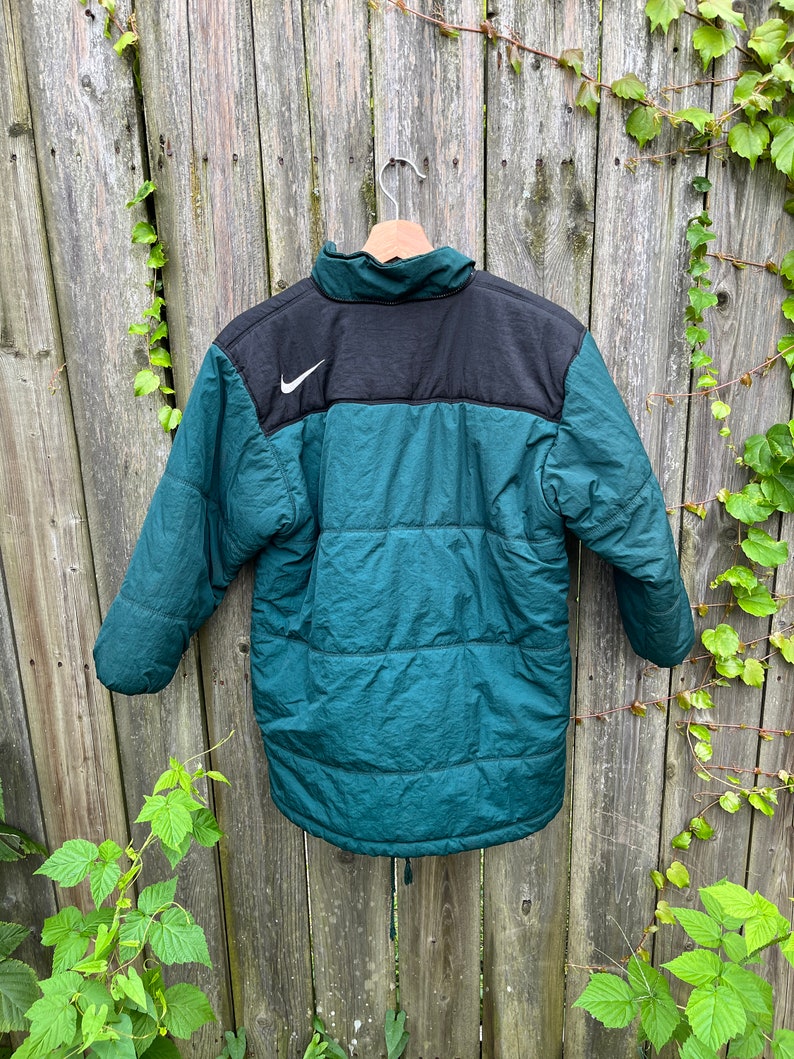 May include: A dark green and black puffer jacket with a white Nike swoosh logo on the upper left chest. The jacket has a zipper closure and a drawstring at the bottom.