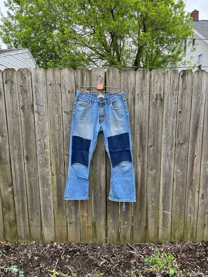 May include: A pair of blue denim bell bottom jeans with dark blue patches on the knees. The jeans are hanging on a wooden fence.