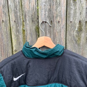 May include: A black and green jacket with a white Nike swoosh logo on the back. The jacket has a zipper closure and a high collar.